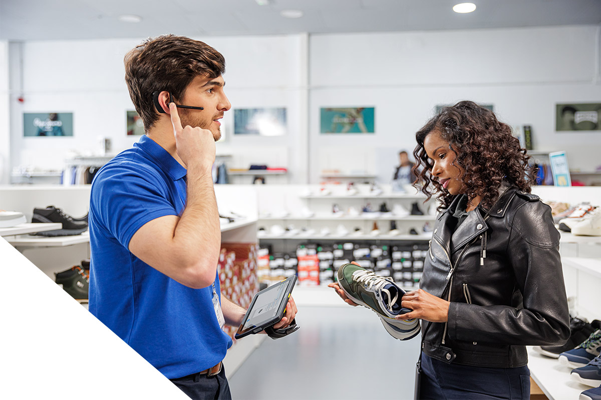 Ein Mitarbeiter im Schuhgeschäft spricht in ein Headset und hält ein Tablet in der Hand. Ihm gegenüber steht eine Frau mit einem Schuh in der Hand.