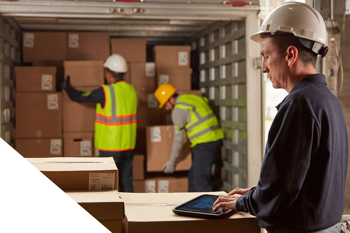 Ein Mann tippt auf einem Tablet, im Hintergrund laden zwei Männer Kartons aus einem Container aus